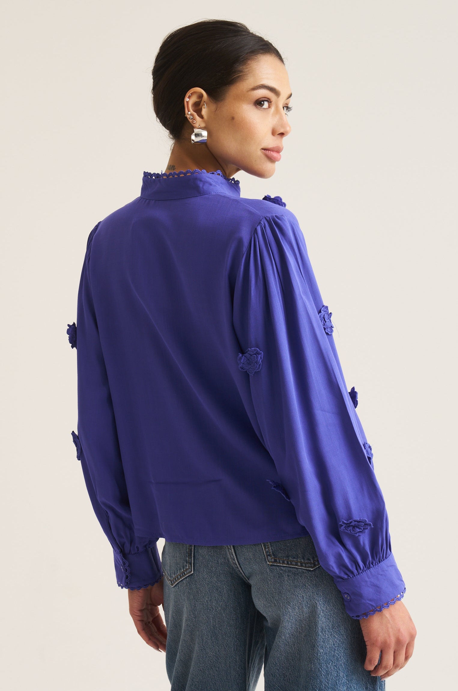 A woman shown from the back wearing a vibrant cobalt long-sleeve blouse with decorative ruffle details on the sleeves and a high ruffled collar, paired with blue jeans, against a plain light beige background.