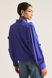 A woman shown from the back wearing a vibrant cobalt long-sleeve blouse with decorative ruffle details on the sleeves and a high ruffled collar, paired with blue jeans, against a plain light beige background.