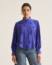 A woman modeling a vibrant Cobalt long-sleeve blouse with 3D floral applique details, paired with wide-leg blue jeans, against a plain light beige studio background.