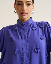 A close-up of a model wearing a vibrant Cobalt long-sleeve blouse with 3D floral appliques and a scalloped button placket.