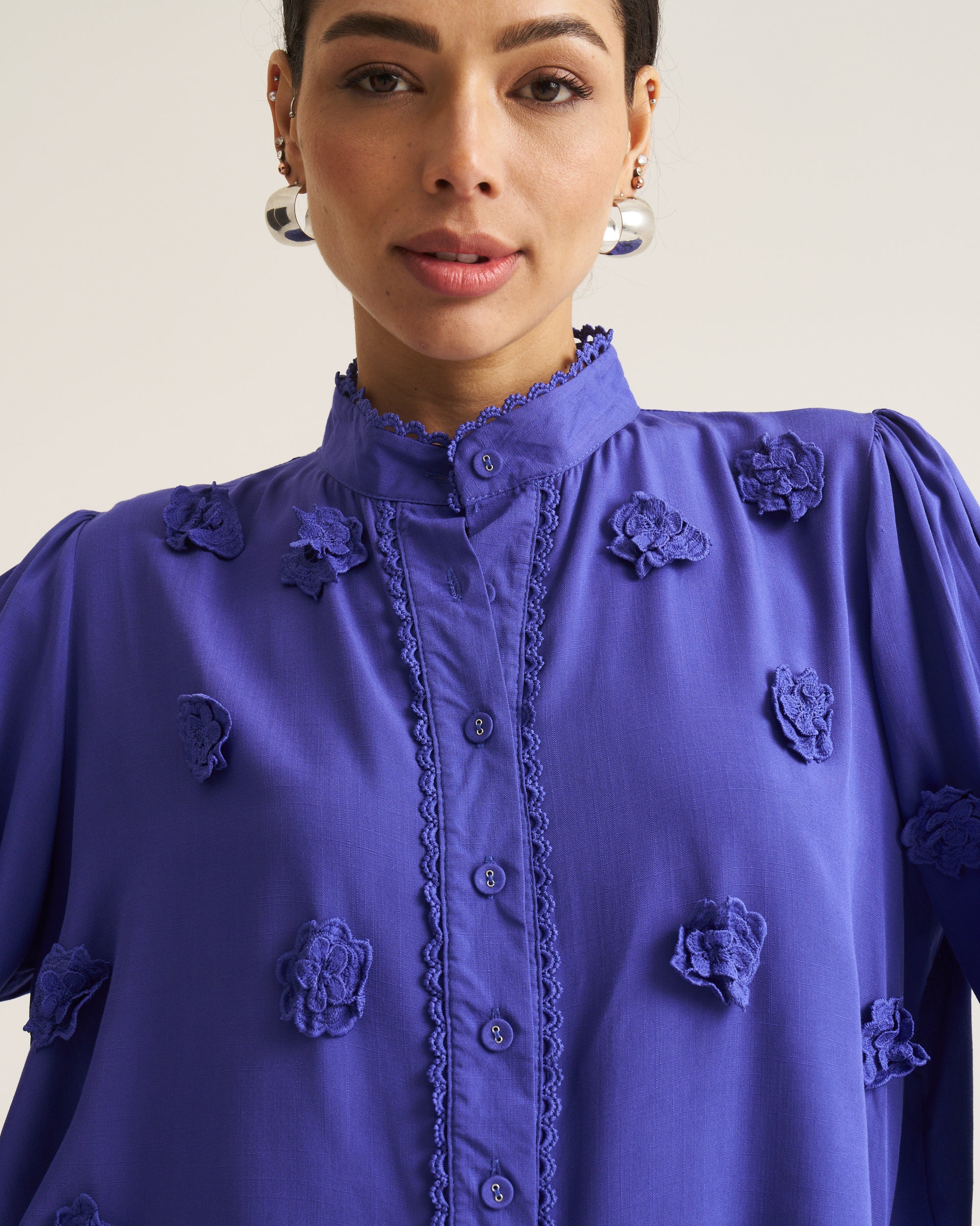 A close-up of a model wearing a vibrant Cobalt long-sleeve blouse with 3D floral appliques and a scalloped button placket.