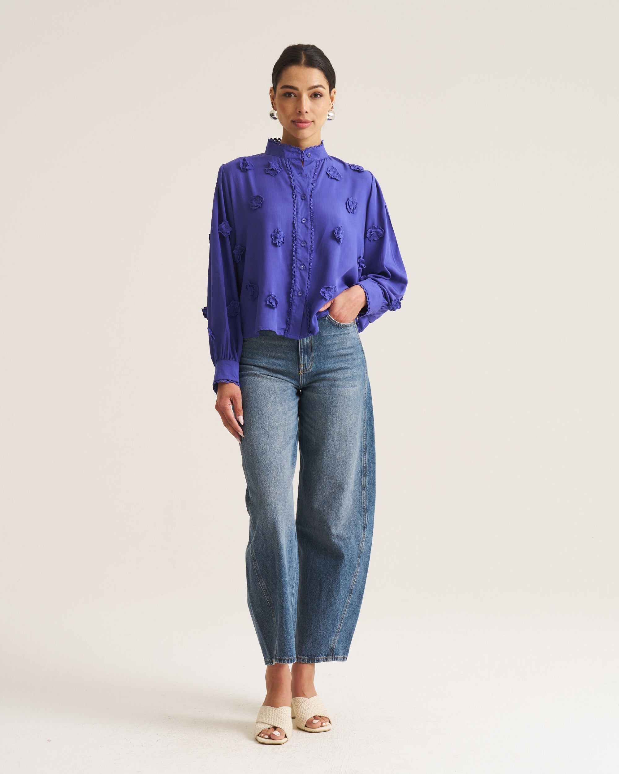 A woman stands against a plain light beige studio background wearing a bright purple embroidered long-sleeve blouse tucked into high-waisted blue jeans with white heeled sandals.
