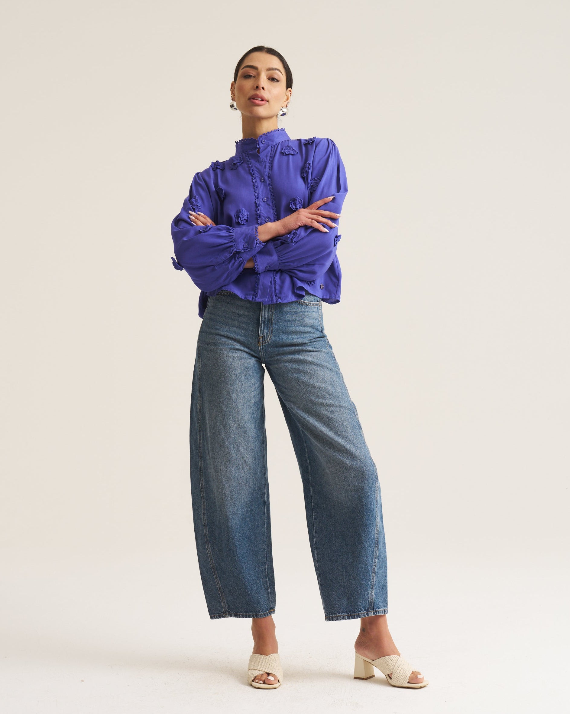 A woman modeling a vibrant cobalt high-neck blouse with dramatic elbow-length sleeves and wide-leg blue jeans, standing against a solid off-white studio background.