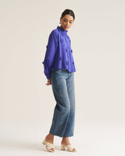 A woman modeling a vibrant cobalt long-sleeve blouse with ruffle details and button accents, paired with blue cropped jeans and beige block-heel sandals, against a plain off-white studio background.