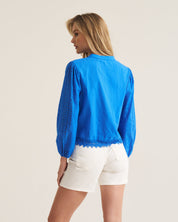 A woman shown from the back wearing a bright blue long-sleeve blouse with lace trim and white shorts, standing against a plain off-white studio background.