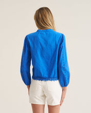 A woman shown from the back wearing a bright blue long-sleeve blouse with lace trim and white shorts, photographed against a plain light beige studio background.