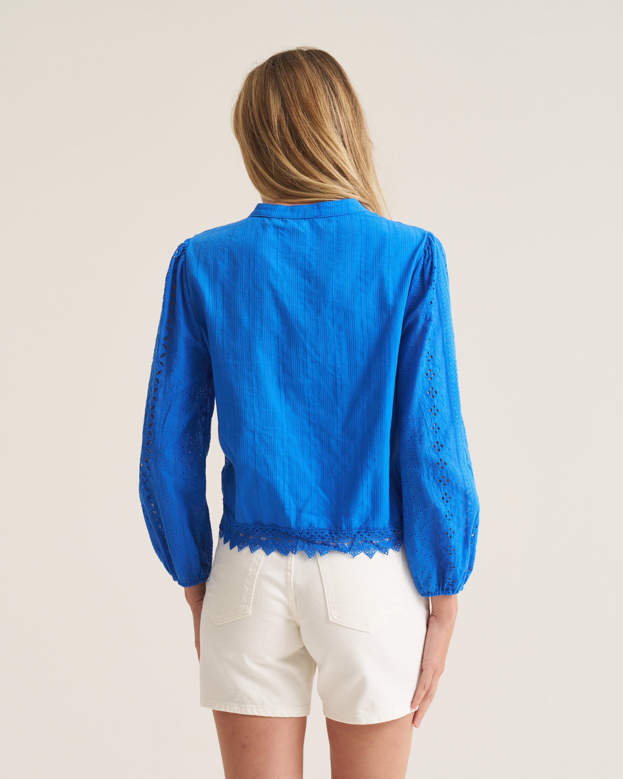 A woman shown from the back wearing a bright blue long-sleeve blouse with lace trim and white shorts, photographed against a plain light beige studio background.