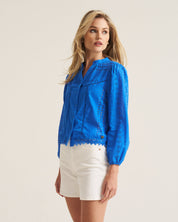 A woman modeling a bright blue eyelet lace blouse with long sleeves and white shorts, standing against a plain light beige studio background.