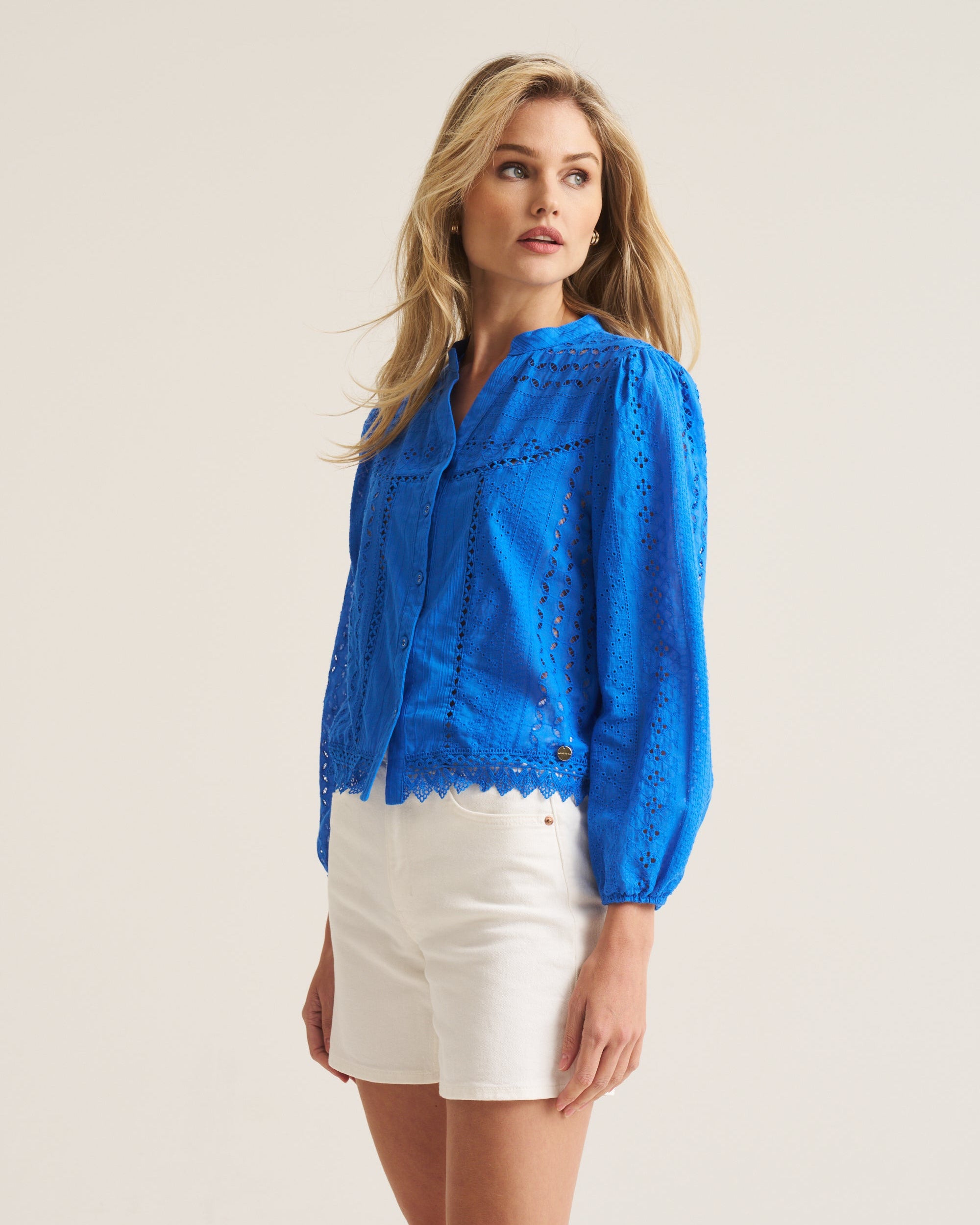 A woman modeling a bright blue eyelet lace blouse with long sleeves and white shorts, standing against a plain light beige studio background.