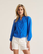 A woman modeling a bright blue long-sleeve eyelet lace blouse with white shorts, standing against a plain light beige studio background.