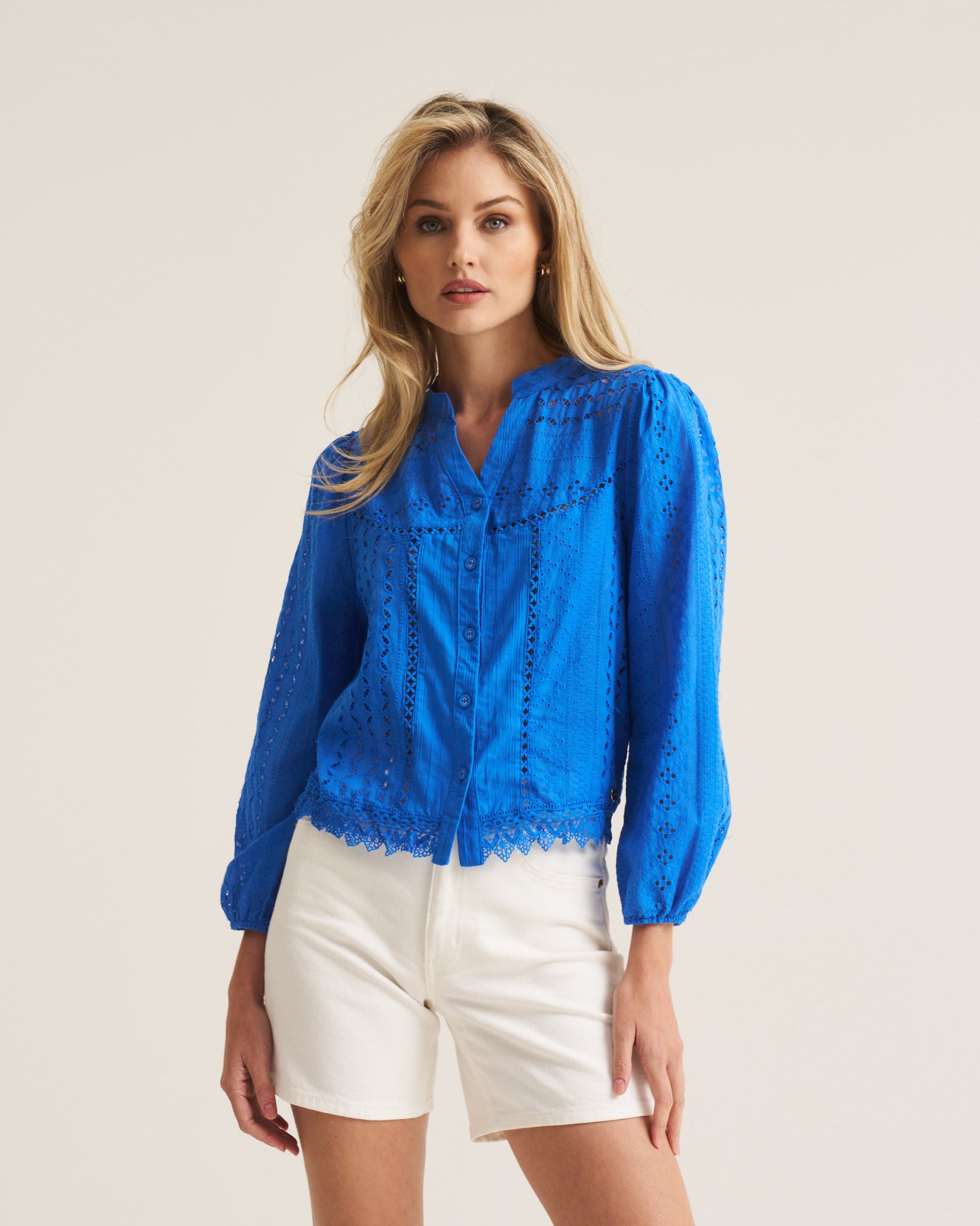 A woman modeling a bright blue long-sleeve eyelet lace blouse with white shorts, standing against a plain light beige studio background.