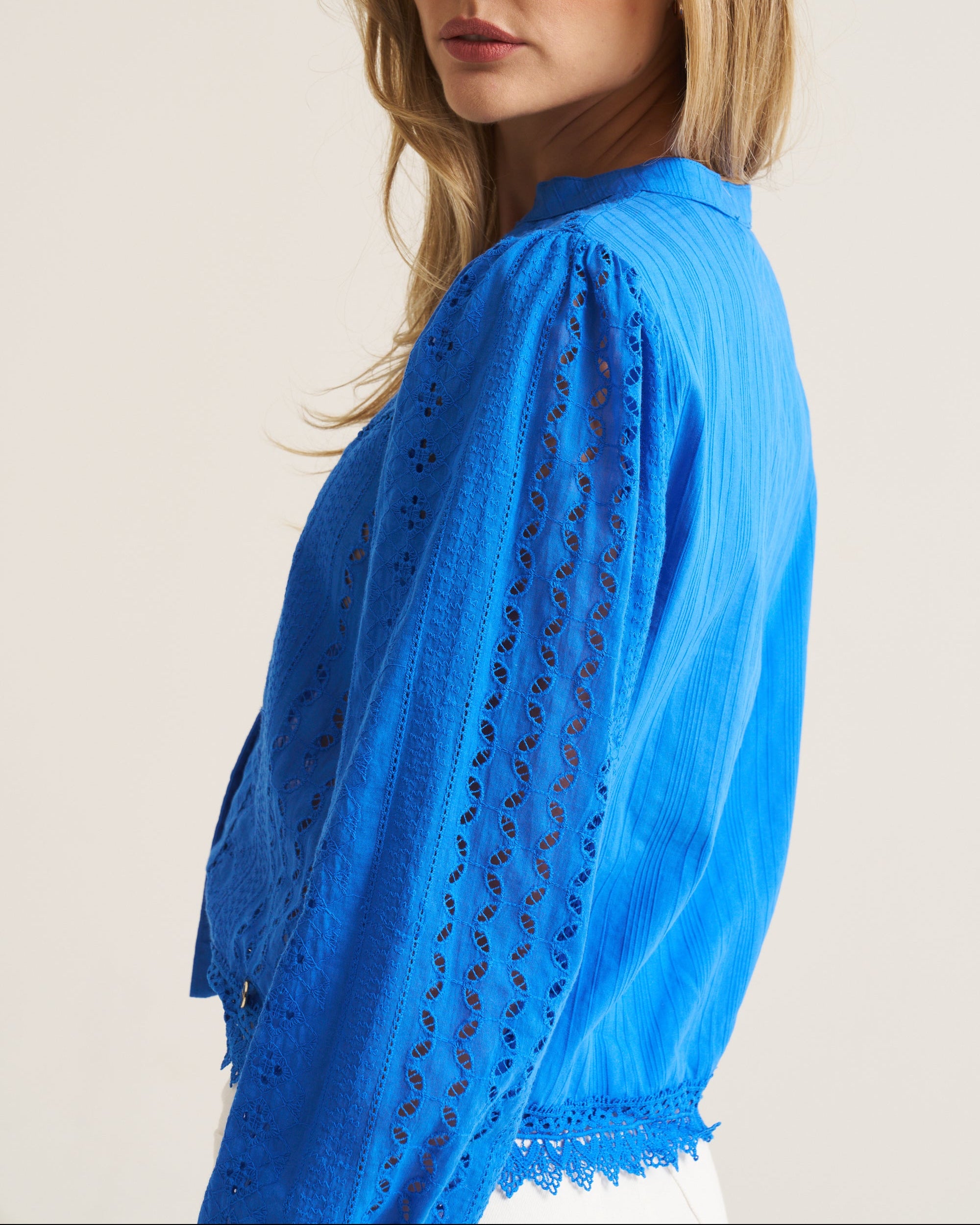 A woman wearing a bright blue eyelet lace blouse, shown from a side-back angle against a plain light beige studio background.