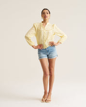 A woman modeling a pale yellow crochet long-sleeve blouse with ruffled details, paired with denim shorts and gold sandals, standing against a plain white studio background.