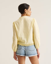 A woman shown from the back wearing a pale yellow long-sleeve blouse with lace-up back detailing and light blue denim shorts against a plain light beige studio background.