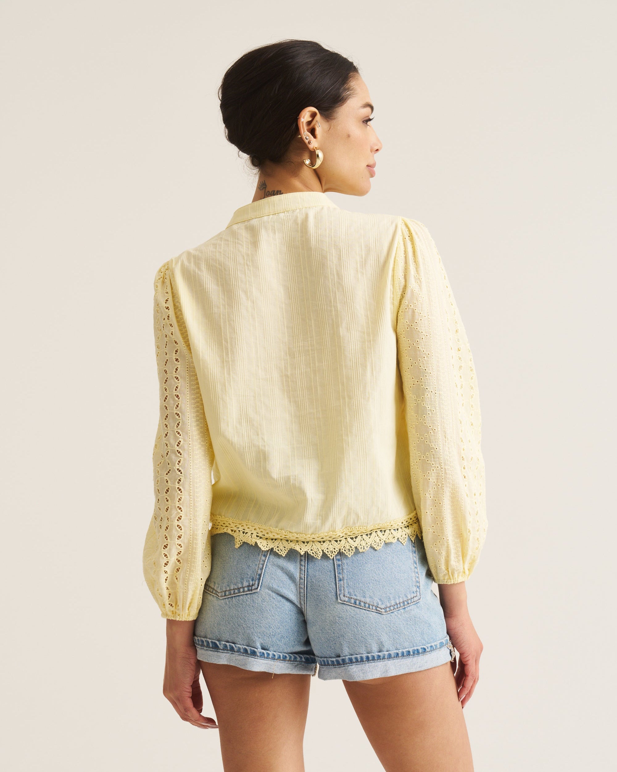 A woman shown from the back wearing a pale yellow long-sleeve blouse with lace-up back detailing and light blue denim shorts against a plain light beige studio background.