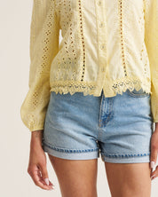 A cropped view of a person wearing a pale yellow eyelet button-up blouse with lace trim and light blue denim shorts against a plain light beige studio background.