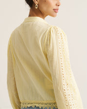A woman shown from the back wearing a pale yellow long-sleeve blouse with lace and eyelet detailing, photographed against a plain light background.