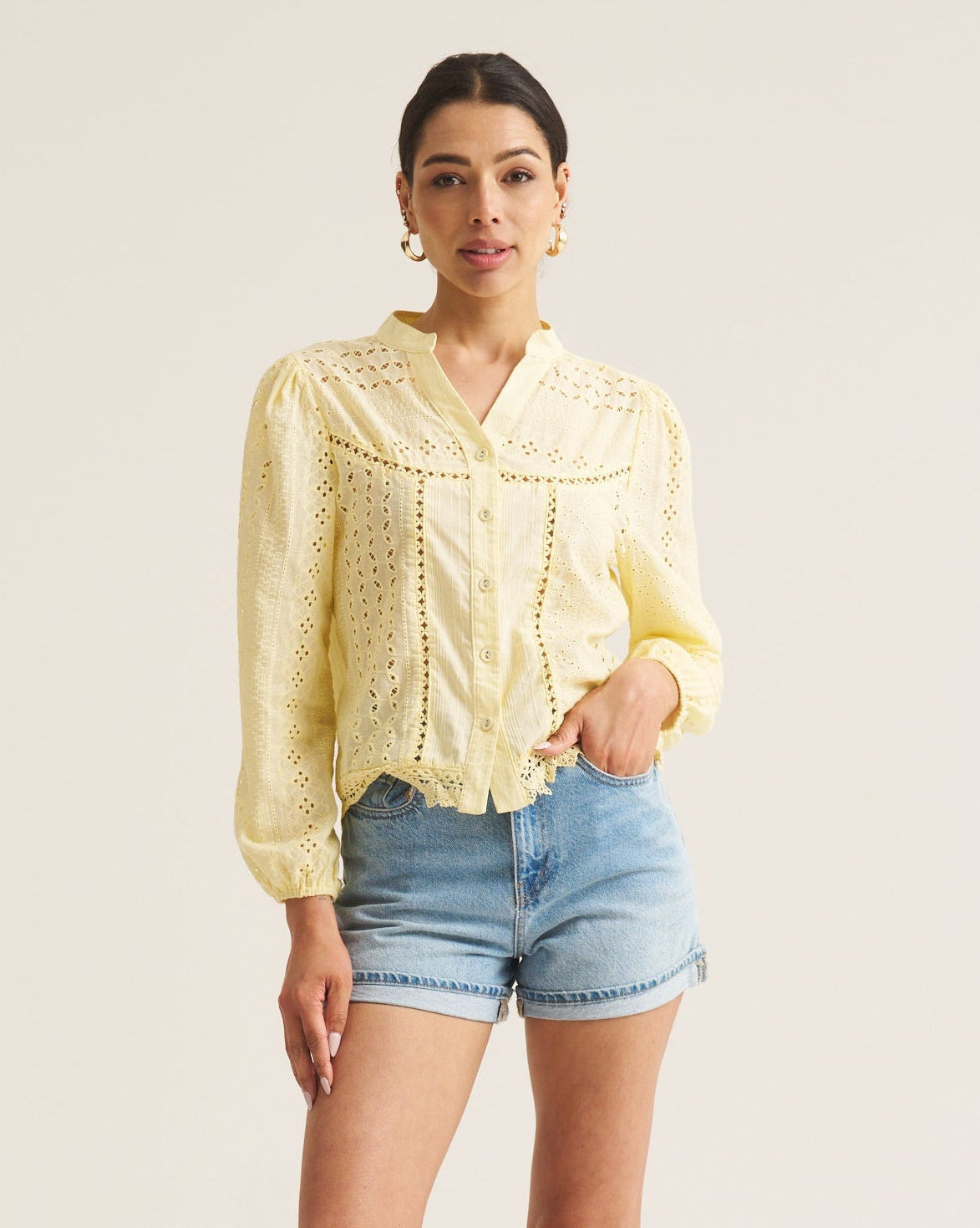 A woman modeling a pale yellow long-sleeve blouse with a textured crochet pattern and denim shorts against a plain off-white studio background.