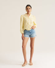 A woman modeling a pale yellow long-sleeve blouse with a textured crochet pattern and denim shorts against a plain off-white studio background.