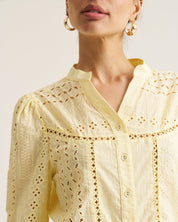 A close-up of a model wearing a pale yellow eyelet blouse with gold embroidery and button details, paired with gold hoop earrings against a light beige background.