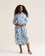 A woman modeling a blue, white, and yellow floral print midi tunic  dress with three-quarter sleeves and a button-down collar, standing against a plain off-white studio background.