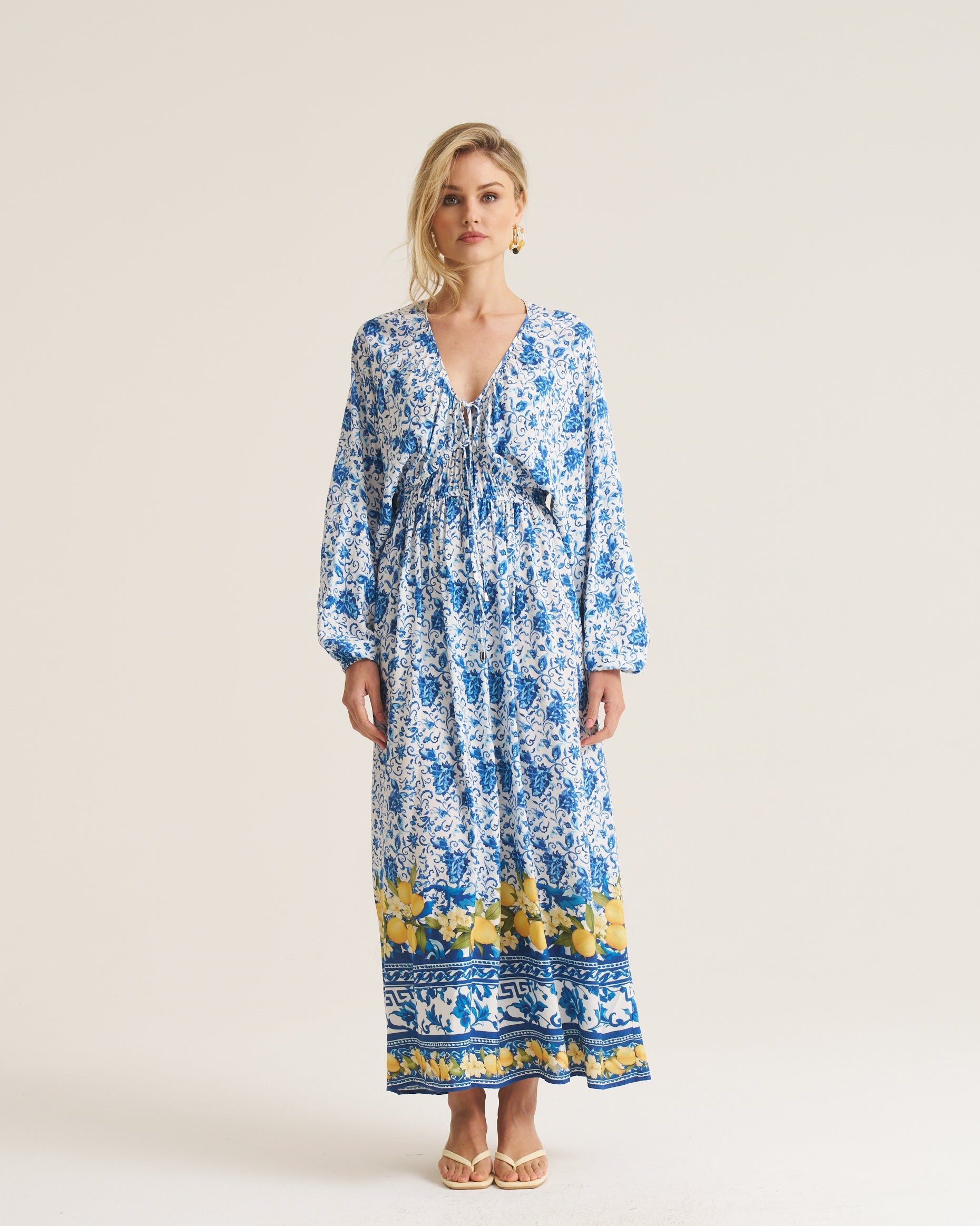 A woman modeling a long-sleeved maxi dress with a blue and white floral print and a yellow patterned border at the hem, standing against a plain off-white studio background.