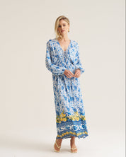 A woman modeling a long-sleeve maxi dress with a blue and white floral pattern and a yellow border print near the hem, standing against a plain light beige studio background.