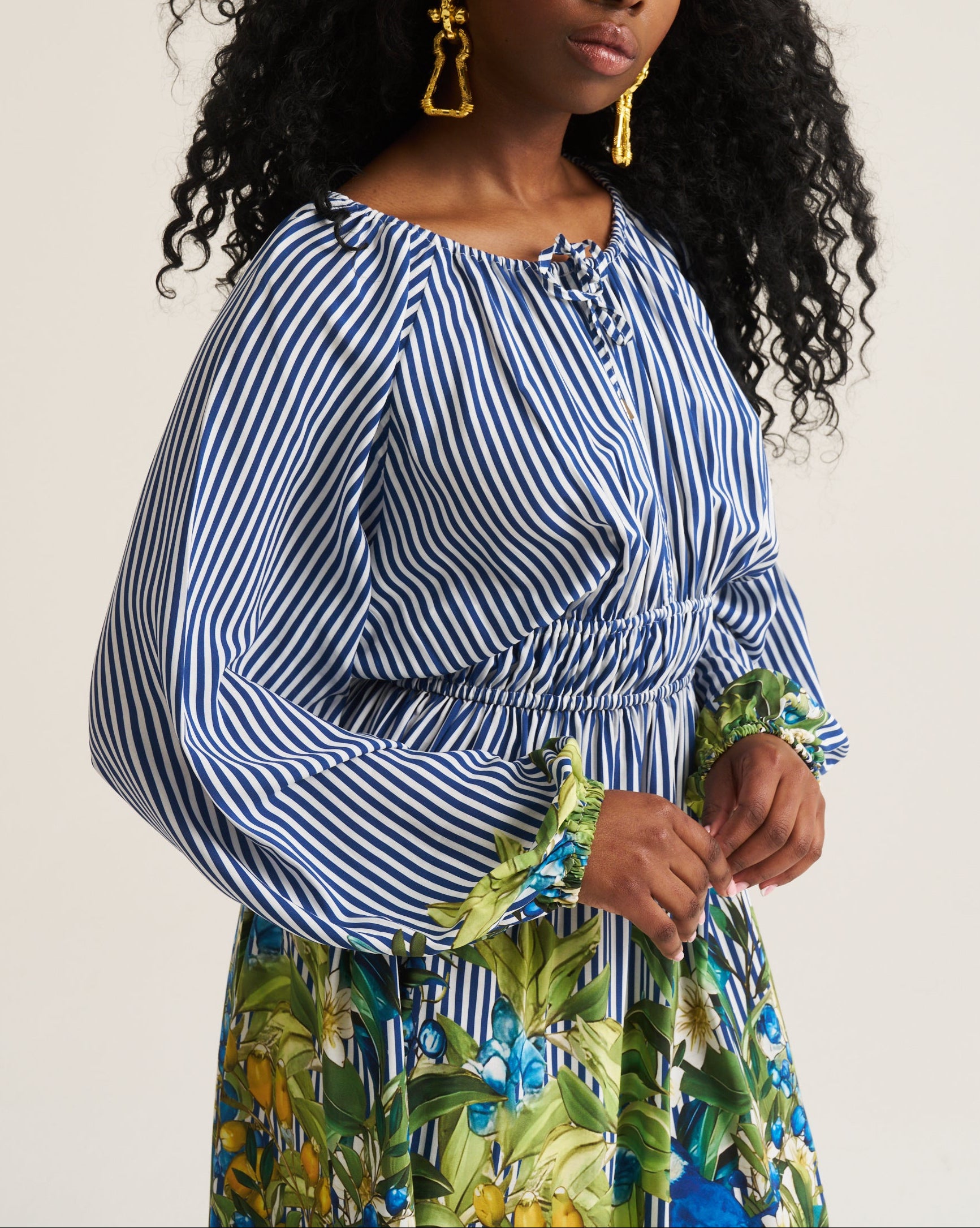 A woman with long, curly black hair is wearing a blue and white vertically striped long-sleeve top with a gathered waist, paired with a green and blue tropical print skirt. She is wearing gold statement earrings and posing against a plain, light beige studio background.