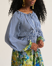 A woman with long, curly black hair is wearing a blue and white vertically striped long-sleeve top with a gathered waist, paired with a green and blue tropical print skirt. She is wearing gold statement earrings and posing against a plain, light beige studio background.