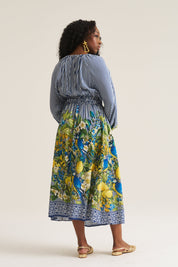 A woman is shown from the back wearing a long-sleeve midi dress with a blue and white striped bodice and a vibrant blue, green, and yellow floral skirt, paired with gold flat sandals against a plain beige studio background.