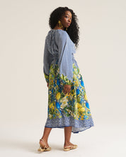 A woman modeling a long-sleeve midi dress with a blue and white striped upper section and a vibrant blue, yellow, and green floral lemon print lower skirt, shown from a three-quarters back view against a plain light beige studio background.