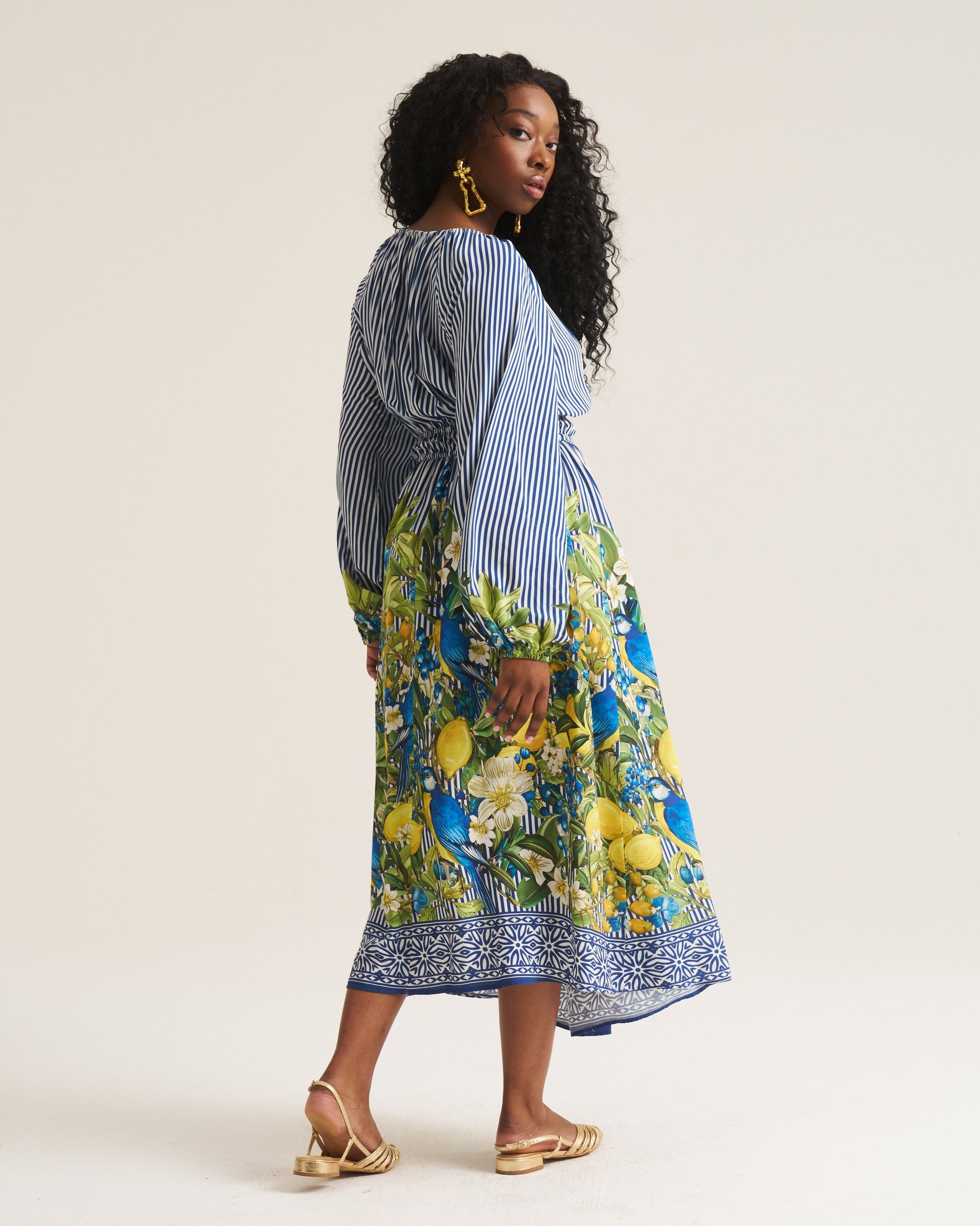 A woman modeling a long-sleeve midi dress with a blue and white striped upper section and a vibrant blue, yellow, and green floral lemon print lower skirt, shown from a three-quarters back view against a plain light beige studio background.