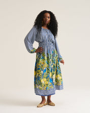 A woman modeling a long-sleeve midi dress with a blue-and-white striped bodice and a vibrant yellow, green, and blue floral print skirt, standing against a plain off-white studio background.