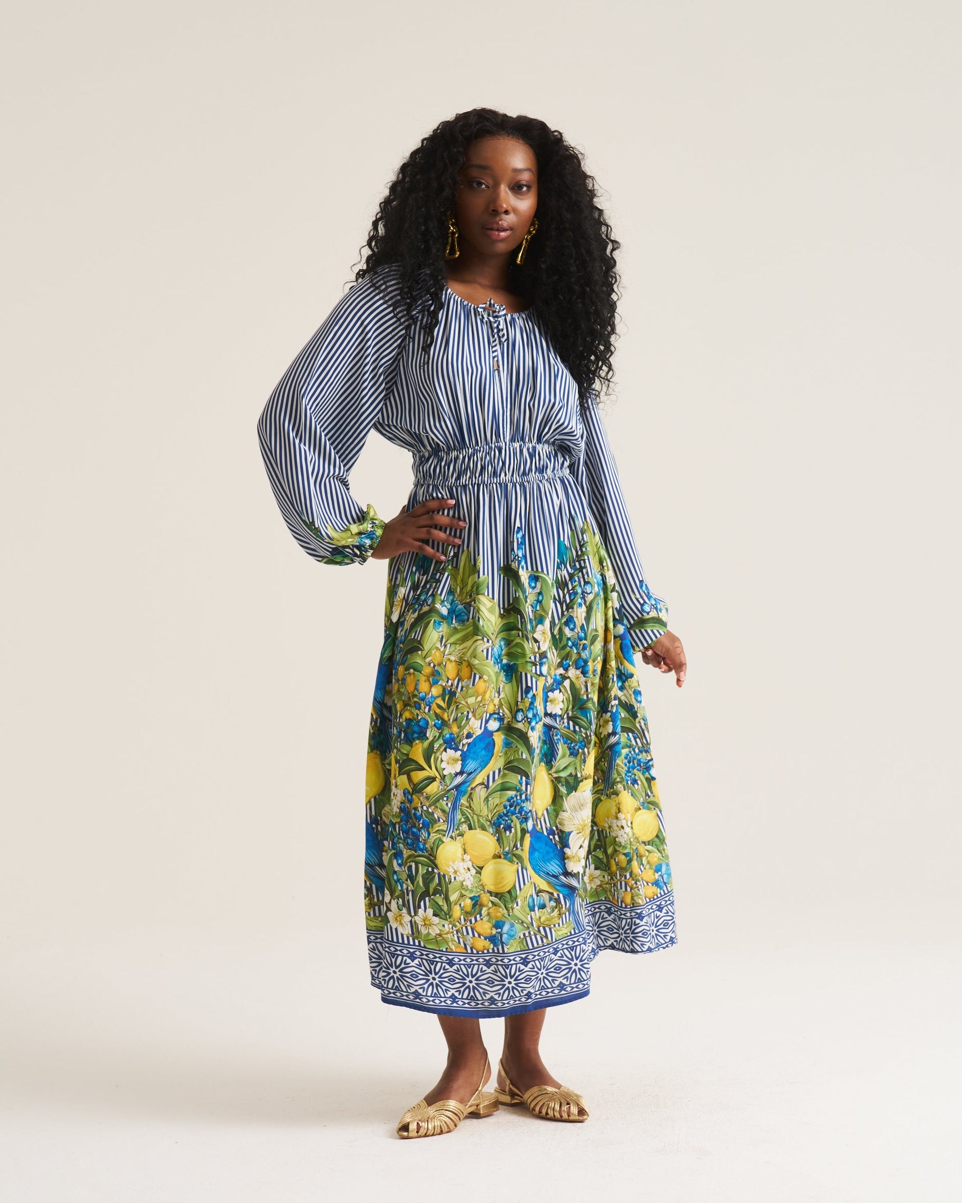 A woman modeling a long-sleeve midi dress with a blue-and-white striped bodice and a vibrant yellow, green, and blue floral print skirt, standing against a plain off-white studio background.