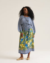 A woman modeling a long-sleeve midi dress with a blue and white striped bodice and a vibrant blue, yellow, and green botanical print skirt, standing against a plain off-white studio background.