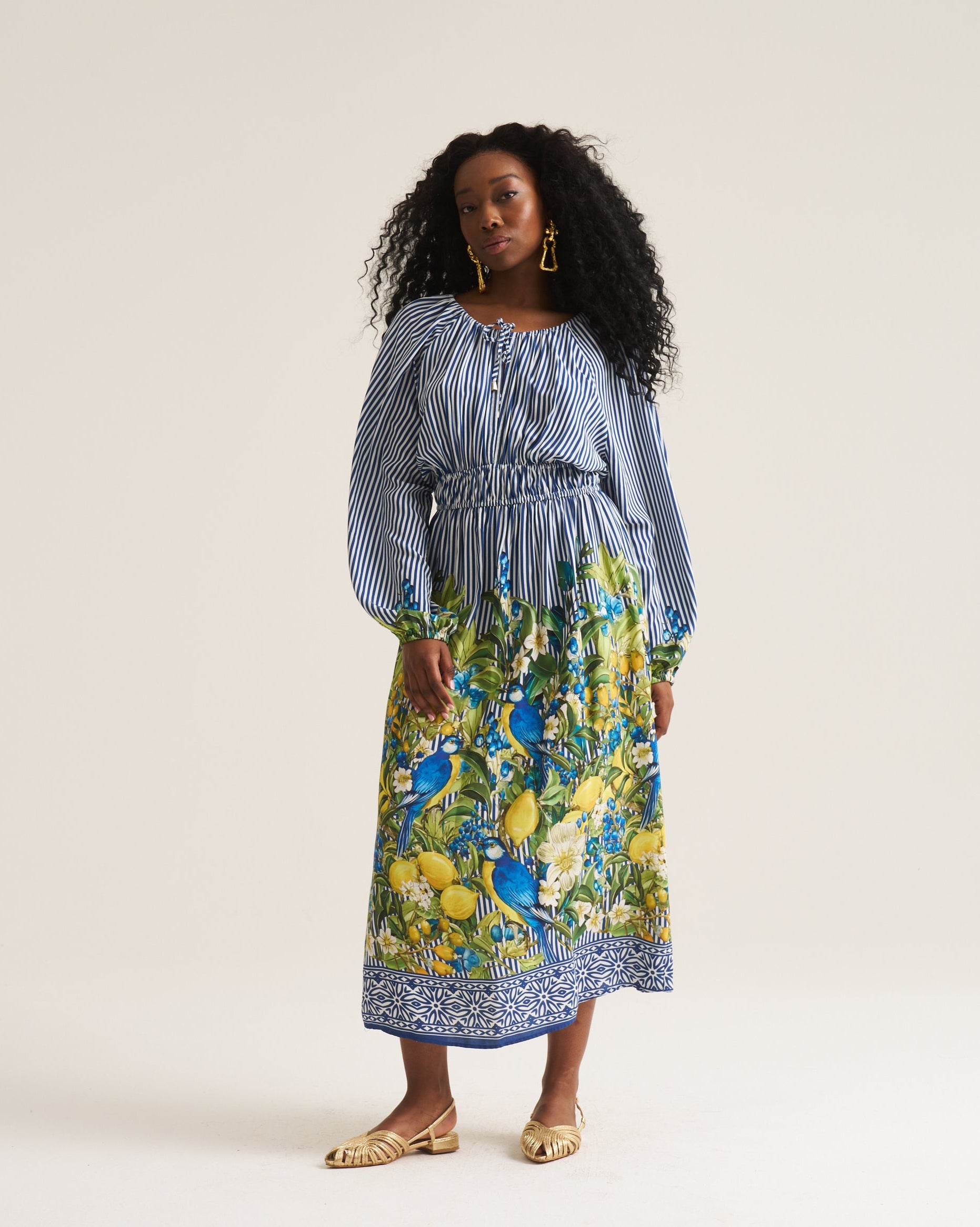 A woman modeling a long-sleeve midi dress with a blue and white striped bodice and a vibrant blue, yellow, and green botanical print skirt, standing against a plain off-white studio background.