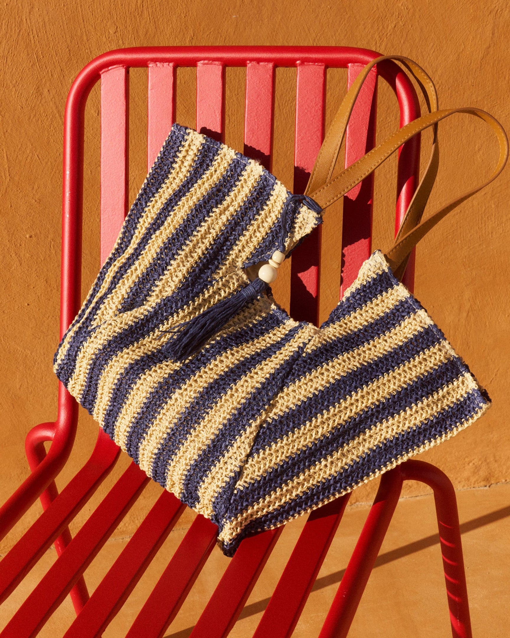 A woven straw tote bag with navy blue and beige horizontal stripes and tan leather handles, resting on a red metal chair against a warm terracotta wall.