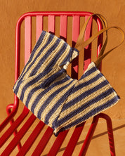 A woven straw tote bag with navy blue and beige horizontal stripes and tan leather handles, resting on a red metal chair against a warm terracotta wall.