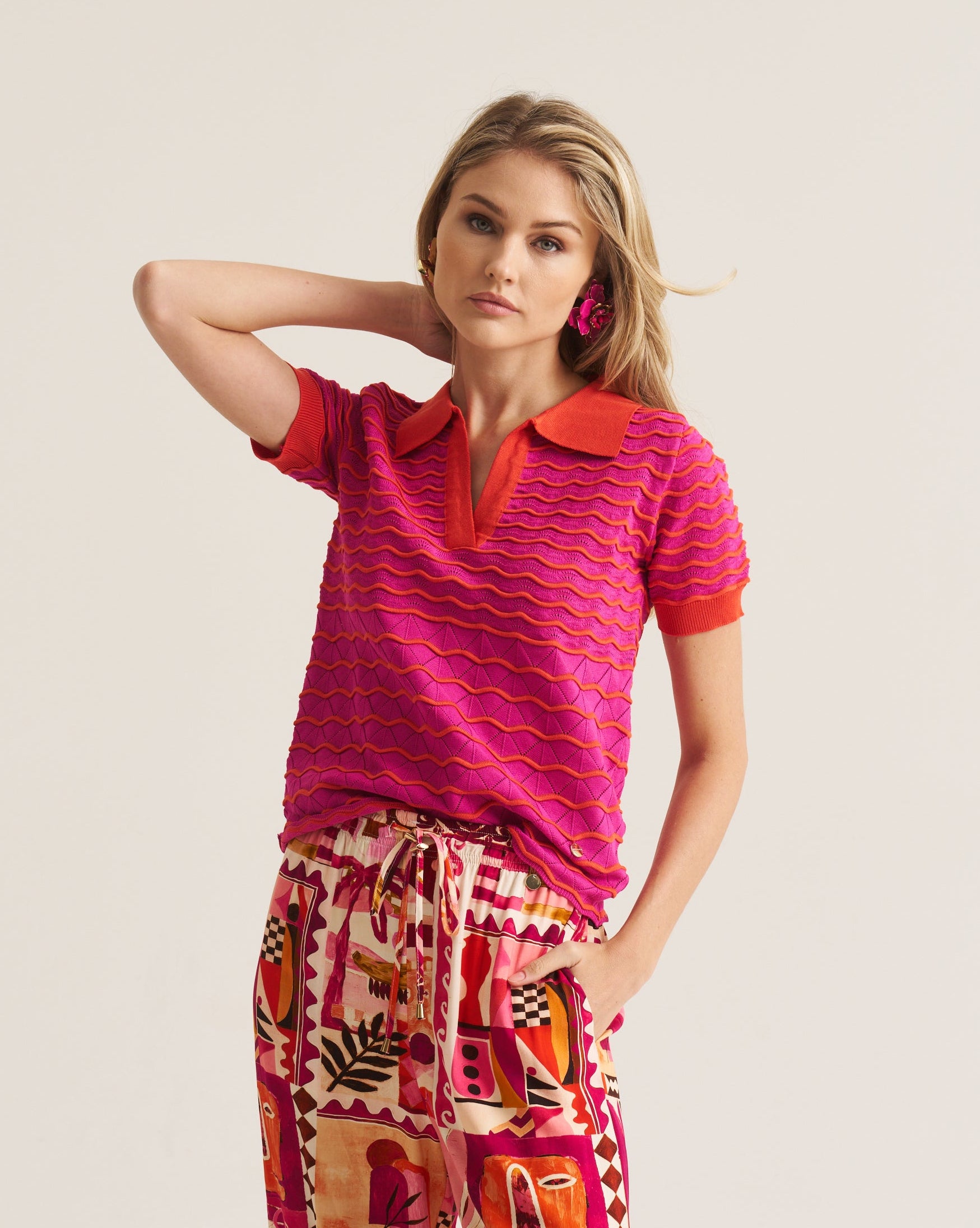 A woman modeling a vibrant pink and red textured short-sleeve polo shirt paired with patterned trousers, set against a plain light beige background.