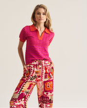 A woman modeling a short-sleeve knit polo shirt in bright pink and orange with matching patterned drawstring pants against a plain light beige studio background.