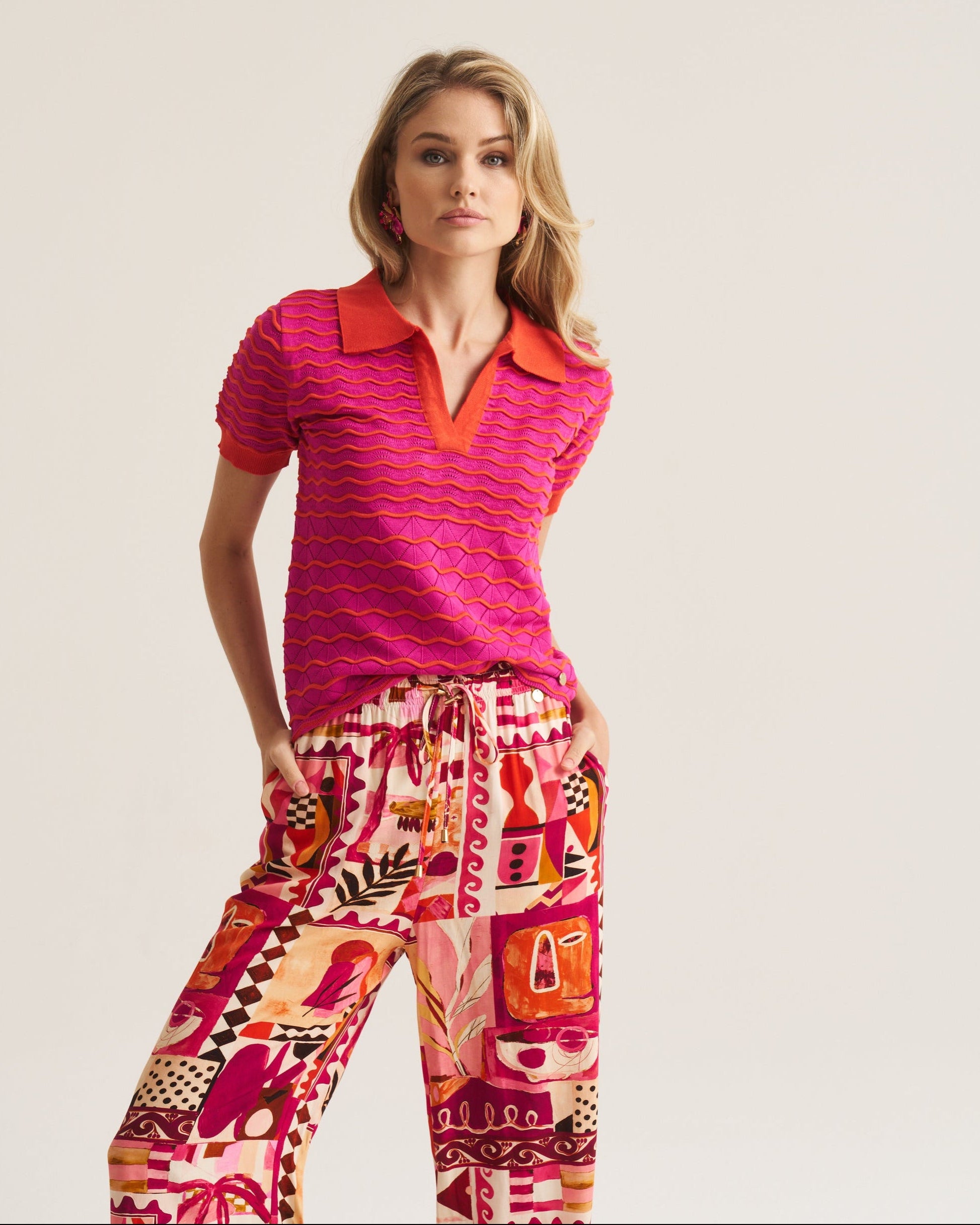 A woman modeling a short-sleeve knit polo shirt in bright pink and orange with matching patterned drawstring pants against a plain light beige studio background.