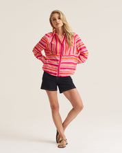 A woman modeling a pink, orange, and yellow striped button-up shirt with black shorts, standing against a plain light beige studio background.