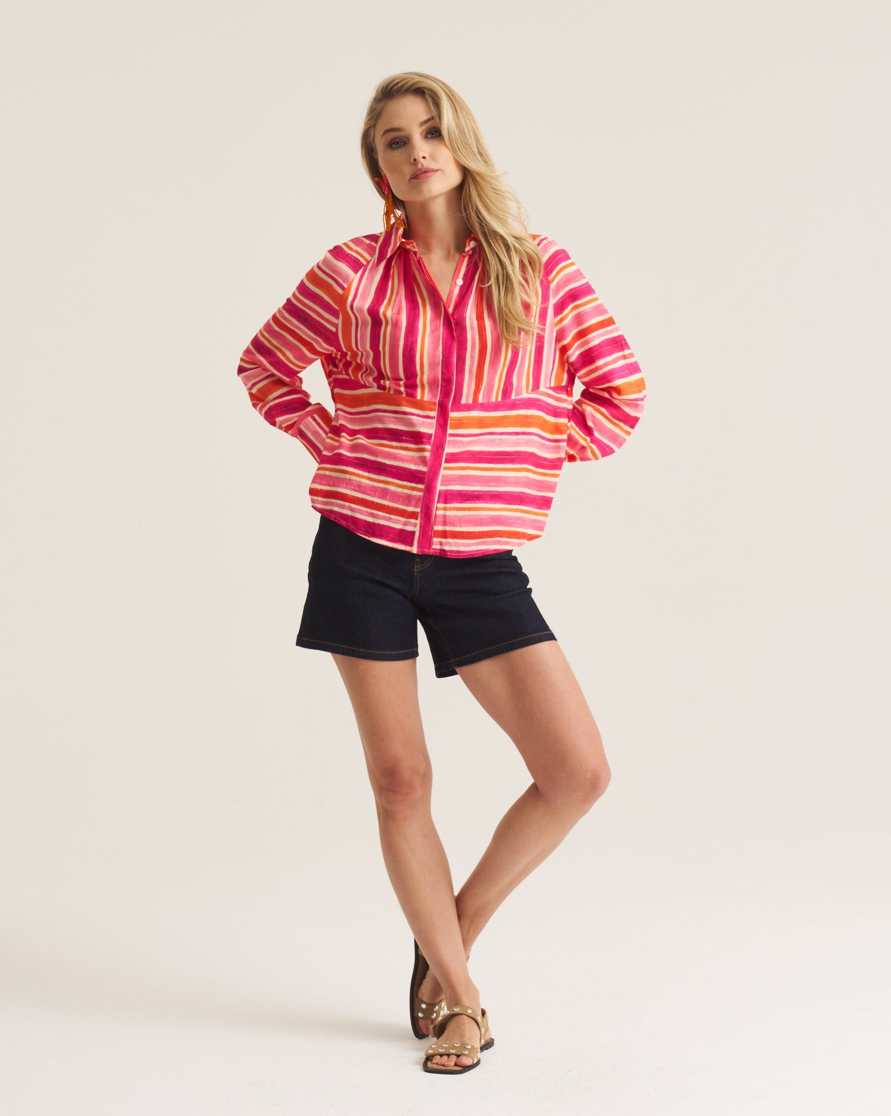 A woman modeling a pink, orange, and yellow striped button-up shirt with black shorts, standing against a plain light beige studio background.