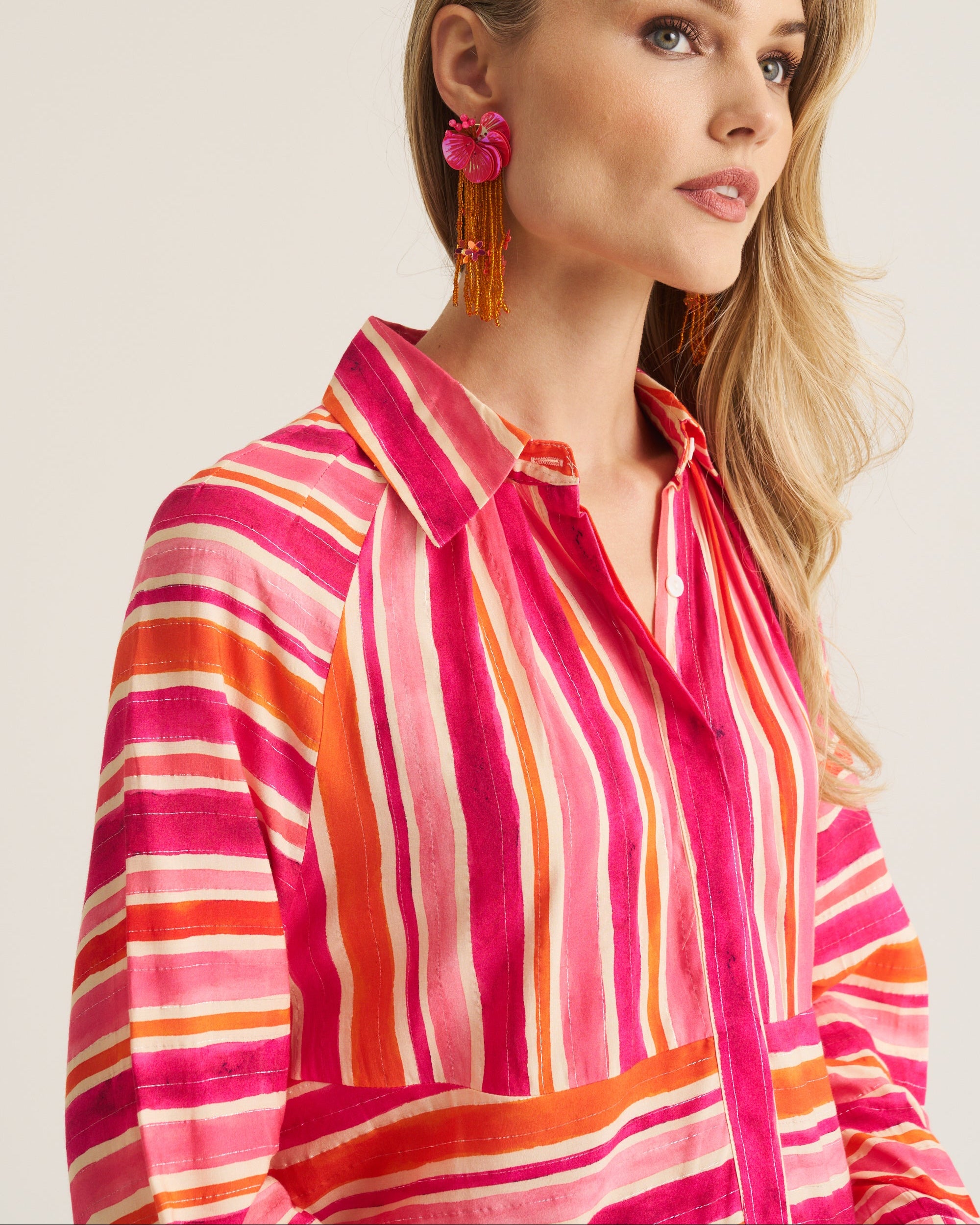 A woman wearing a vibrant pink, orange, and white striped button-up shirt, paired with bright pink and orange tassel earrings, set against a plain light beige background.