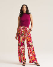 A woman modeling a sleeveless magenta top and high-waisted, wide-leg trousers featuring a vibrant, multicolored patchwork print in pink, orange, red, and cream. She is standing against a plain, light beige studio background.