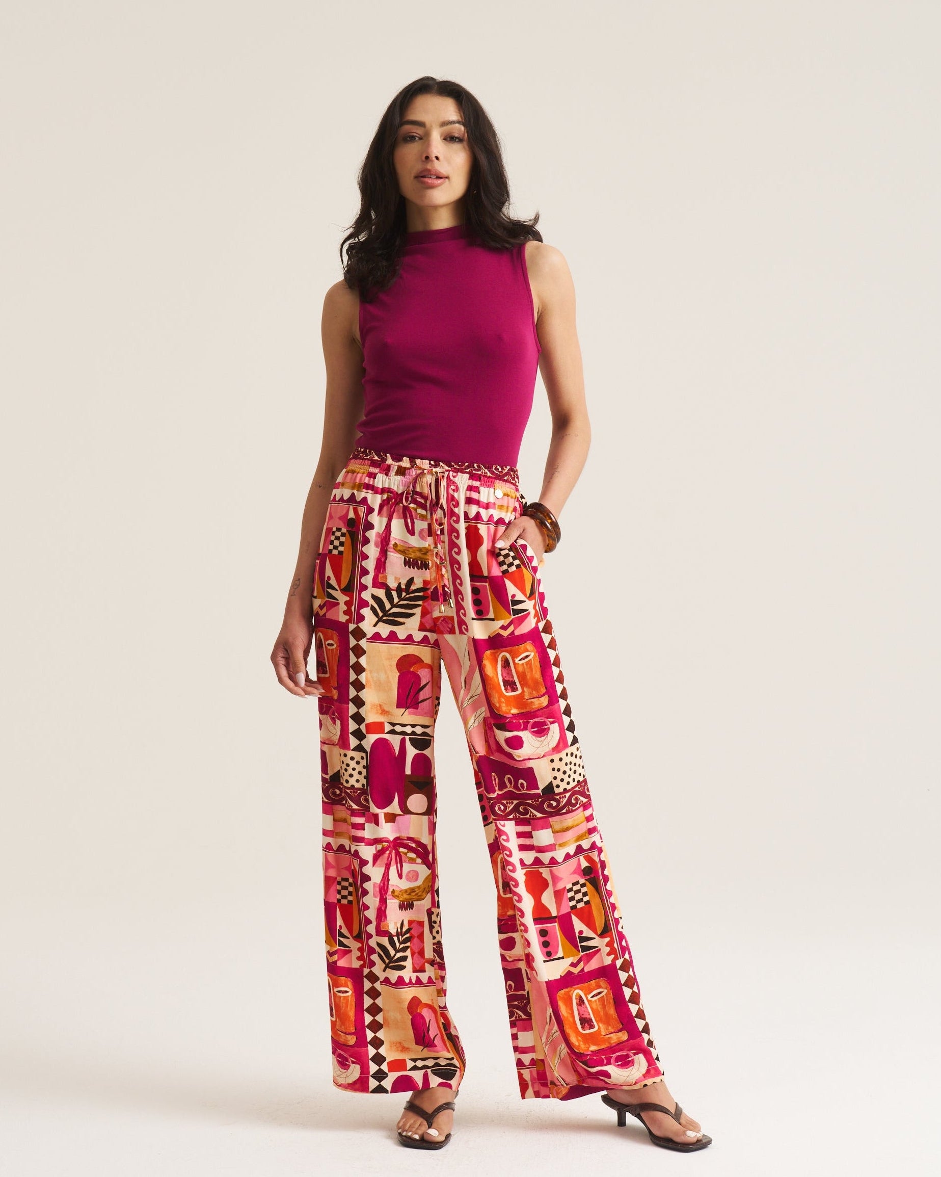A woman modeling a sleeveless magenta top and high-waisted, wide-leg trousers featuring a vibrant, multicolored patchwork print in pink, orange, red, and cream. She is standing against a plain, light beige studio background.