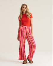 A woman modeling a bright red short-sleeve top and high-waisted, wide-leg trousers with vertical pink, red, and orange stripes, paired with brown sandals, against a solid off-white studio background.