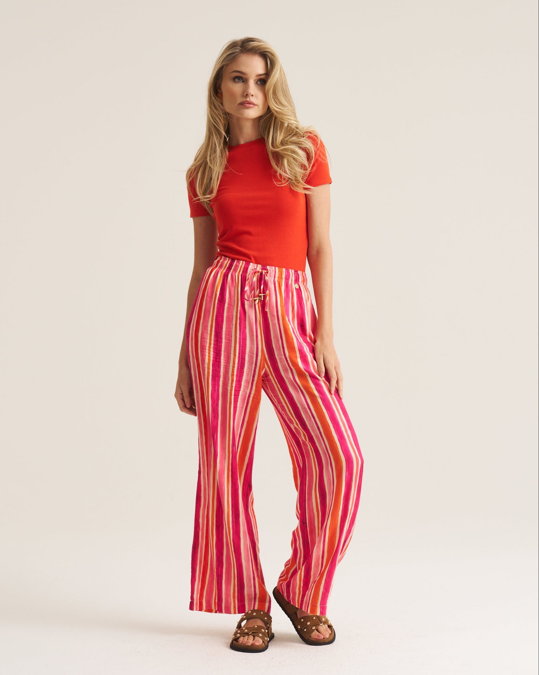 A woman modeling a bright red short-sleeve top and high-waisted, wide-leg trousers with vertical pink, red, and orange stripes, paired with brown sandals, against a solid off-white studio background.