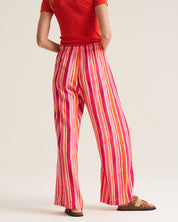 A model shown from the back wearing wide-leg trousers with vertical pink, orange, yellow, and white stripes, paired with a red t-shirt and brown slide sandals, against a plain light beige background.