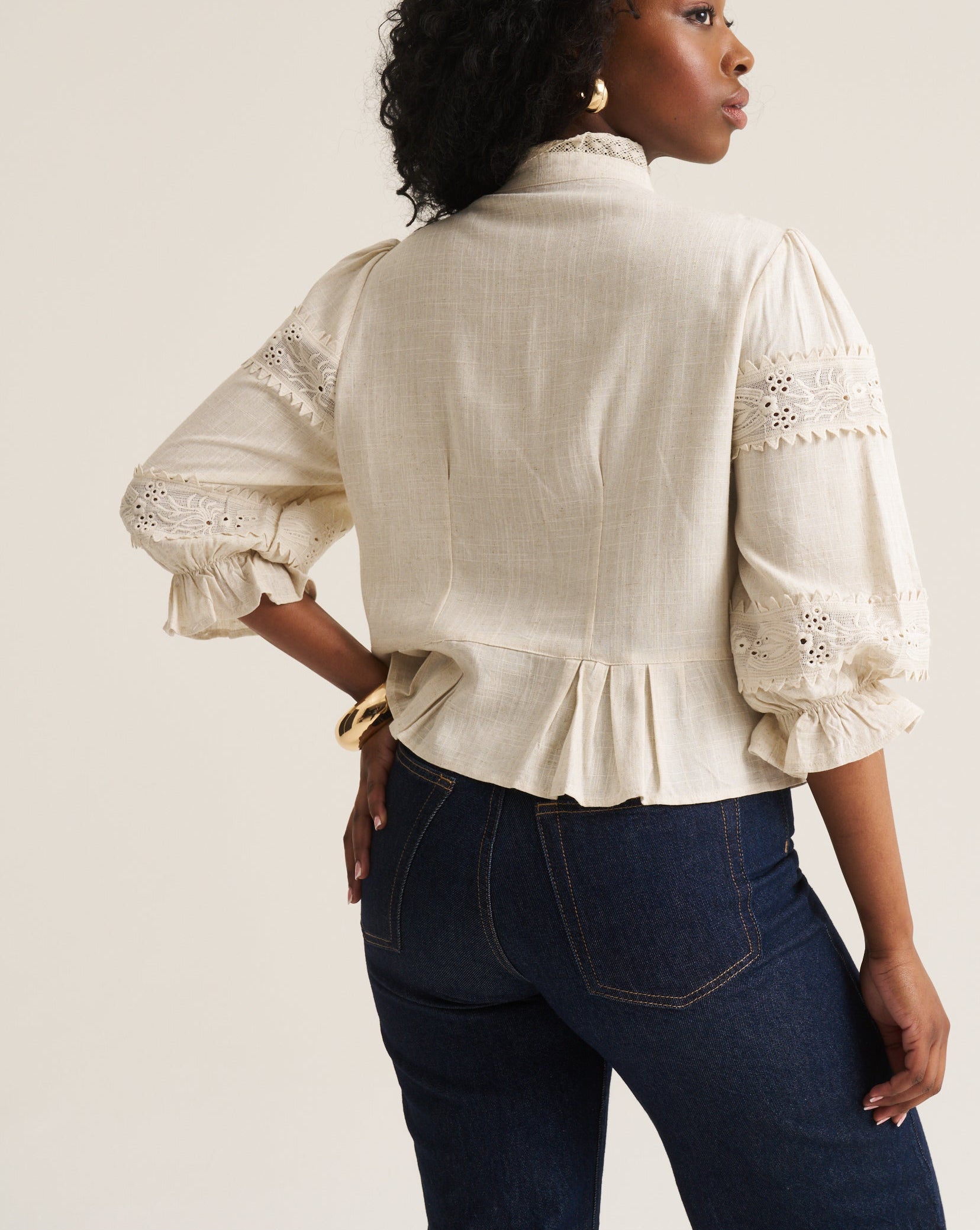 A woman shown from the back wearing a cream-colored blouse with lace sleeve details and dark blue jeans, standing against a plain off-white studio background.